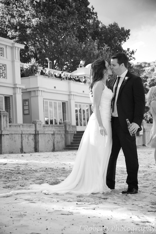 Bride and groom kissing on Balmoral Beach with guests on Bather's Pavilion behind them - wedding photography sydney